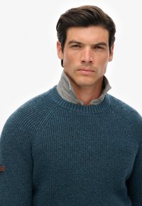 Blauer gerippter Pullover mit einem grauen Kragen, runder Ausschnitt und Raglanärmeln. Verfügt über dezente Leder-Logos auf dem linken Arm.