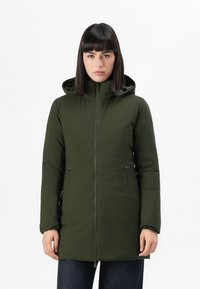 Vrouw in een olijfgroene, met rits gesloten winterjas met capuchon, met ontspannen handen aan de zijkanten tegen een effen witte achtergrond.