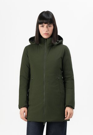 Vrouw in een olijfgroene, met rits gesloten winterjas met capuchon, met ontspannen handen aan de zijkanten tegen een effen witte achtergrond.