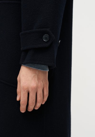 Χέρι που ακουμπάει δίπλα σε μανίκι σκούρου μπλε παλτού με ιμάντα με κουμπί πάνω από μανσέτα γκρι πουλόβερ μπροστά σε απλό φόντο.