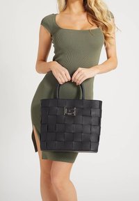 Sac à dos tissé noir avec double poignées, mettant en avant un accent logo doré. Fabriqué en matériau texturé, il a une forme rectangulaire.