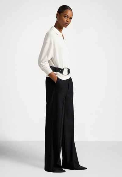 Mulher está em pé com as mãos nos bolsos, vestindo uma camisa polo branca de manga comprida, calças largas pretas de cintura alta e um cinto preto grande.