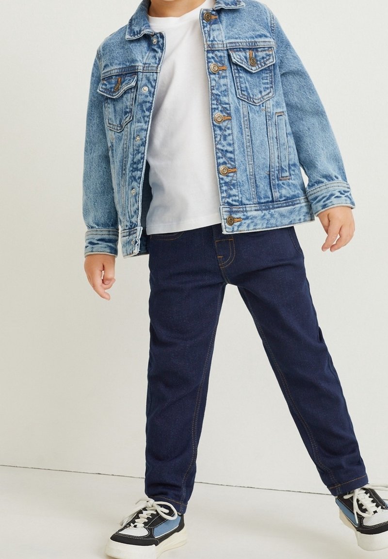 Enfant portant une veste en jean bleu clair, un t-shirt blanc, un jean bleu foncé et des baskets noires et blanches, debout, jambes écartées.