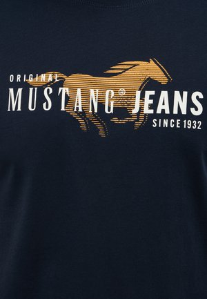 Námorníckom modrom bavlnenom tričku s grafikou zlatého koňa a bielym textom „Originálne Mustang Jeans od roku 1932.“ Hladký povrch.