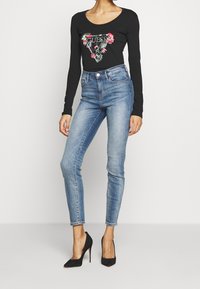 Top nero a maniche lunghe con stampa floreale e logo, abbinato a jeans skinny azzurri chiari e tacchi a spillo neri. Design semplice e aderente.