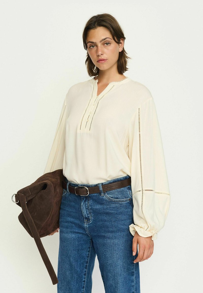 Krämfärgad blus med henleykrage, långa puffärmar, knappslå och spetsdetaljer. Matchas med blå jeans och en brun väska.