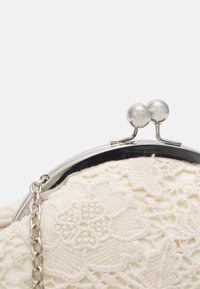 Kermainen pitsikäsipurje hopeisella metalliremmillä ja nepparilla, jossa kukkaisemerkki ja ketjuhihna. Näkyy teksturoitu pinta.