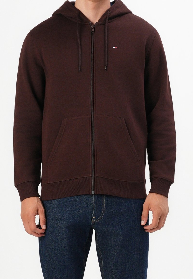 Homme portant un sweat à capuche marron foncé zippé avec des poches avant et un jean bleu, mains détendues le long du corps, sur un fond uni.