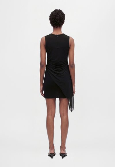 Modèle vu de dos portant une mini-robe noire sans manches, avec des fronces drapées sur les côtés et un panneau transparent retombant, assortie à des talons noirs.