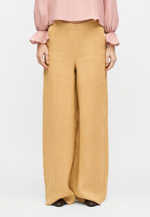 Žena oblečená v béžových širokých nohaviciach s vysokým pásom, doplnených ružovou blúzkou s dlhými rukávmi a tmavými topánkami na bielom pozadí.