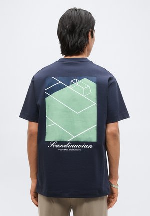 Isik, kes kannab tumesinist T-särki, millel on roheline ja valge jalgpalliväljaku graafika ning tagaküljel tekst "Scandinavian Football Community".