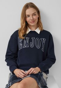 Sudadera navy con el texto blanco "ENJOY", con cuello redondo y mangas largas, combinada con una falda de cuadros en tonos azules y grises.