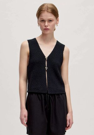 Zwart mouwloos topje van een gestructureerde stof, met V-hals en een rits aan de voorkant met gouden hartvormige hardware. Gestyled met zwarte shorts.