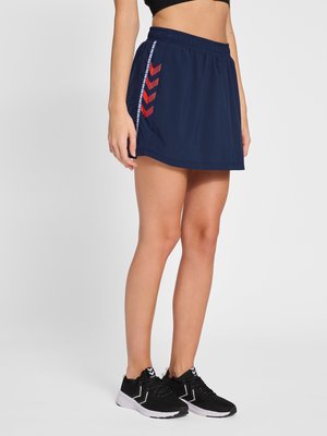 Vrouw draagt een marineblauwe sportrok met rode chevronstrepen en zwarte hardloopschoenen op een witte achtergrond.