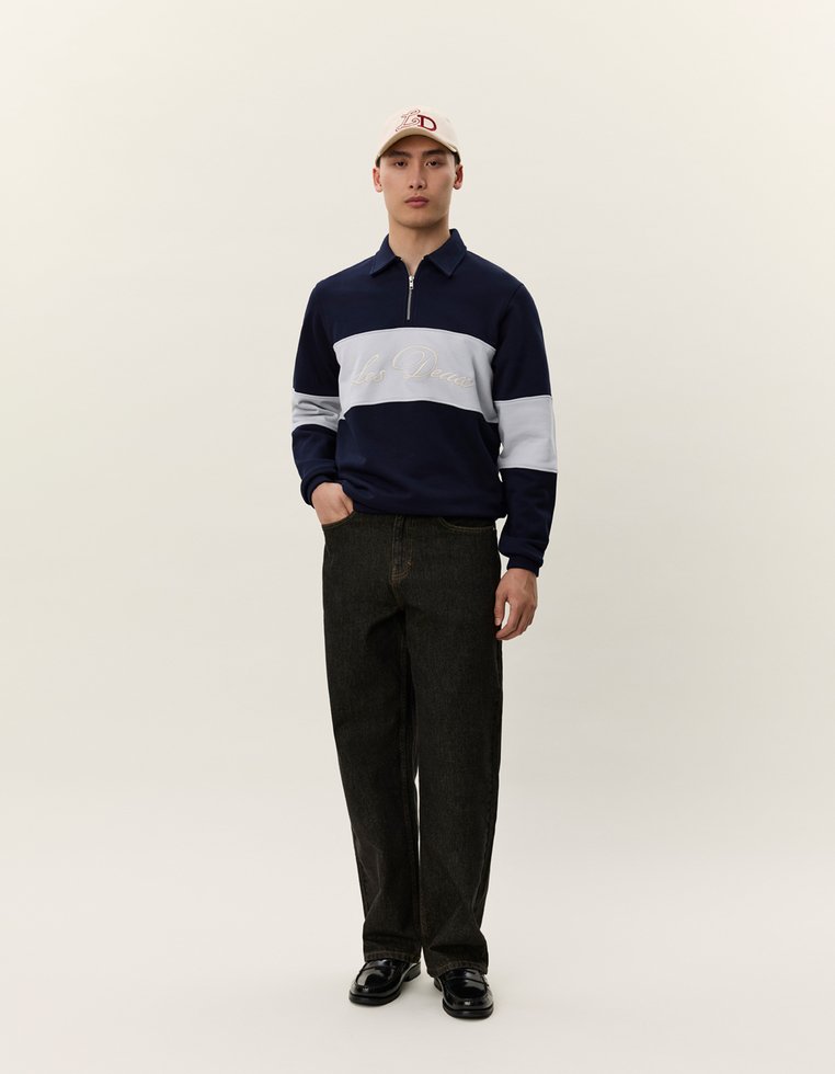 Námornícke a svetlosivé zip-up pulóver s golierom, s textom. Čierne široké nohavice a svetlá čiapka dotvárajú outfit.
