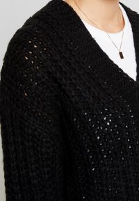 Svart stickad kardigan med lös, öppen väv och bred krage; bärs över en vit skjorta med ett litet rektangulärt guldhänge.