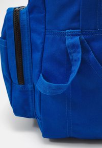 Modrý textilní batoh s texturovaným povrchem, boční kapsou a černým zipem. Obsahuje robustní rukojeť po straně.