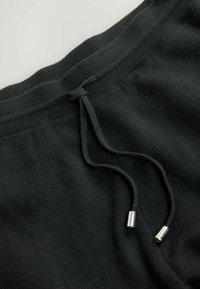 Schwarze Jogginghosen aus weichem Stoff mit einem gerippten Bund und einem Kordelzug mit metallischen Aglets. Die Oberfläche ist glatt mit leichter Struktur.