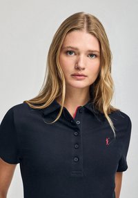 Marinblå polotröja med krage, som har en femknapplång öppning. Utmärkande röd logotyp accent på bröstet. Slät, texturerad tyg.