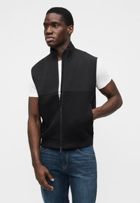 Μαύρο γιλέκο χωρίς μανίκια με ψηλό γιακά, φερμουάρ μπροστά, πάνελ υφάσματος σε αντίθετο χρώμα, μαλακή υφή, φορεμένο πάνω από λευκό μπλουζάκι και μπλε τζιν.
