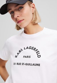 Balta marškinėliai su ryškiu juodu tekstu, skaitančiu „Karl Lagerfeld Paris 21 Rue St-Guillaume“. Trumpi rankovės ir apvalus kaklas.