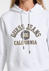Valkoinen huppari, jossa on etupuolella grafiikka, jossa lukee "Guess Jeans California." Hupparissa on kengurutasku, nyörit, ribattu hihansuu ja pehmeä, teksturoitu kangas.