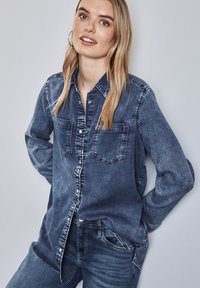 La camicia in denim di blu sbiadito presenta maniche lunghe, una chiusura frontale con bottoni e due tasche sul petto. Il tessuto ha una consistenza morbida e usurata con cuciture a contrasto.