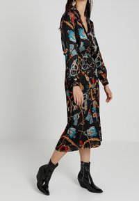 Vestido midi negro con vibrantes patrones florales y de cadenas. Mangas largas, cuello y cintura ceñida. La modelo lo combinó con botines negros brillantes.