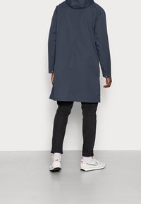 Person som bär en marinblå huvjacka, svarta uppvikta byxor, vita strumpor och vita Nike-sneakers, stående på ett grått golv mot en vit vägg.