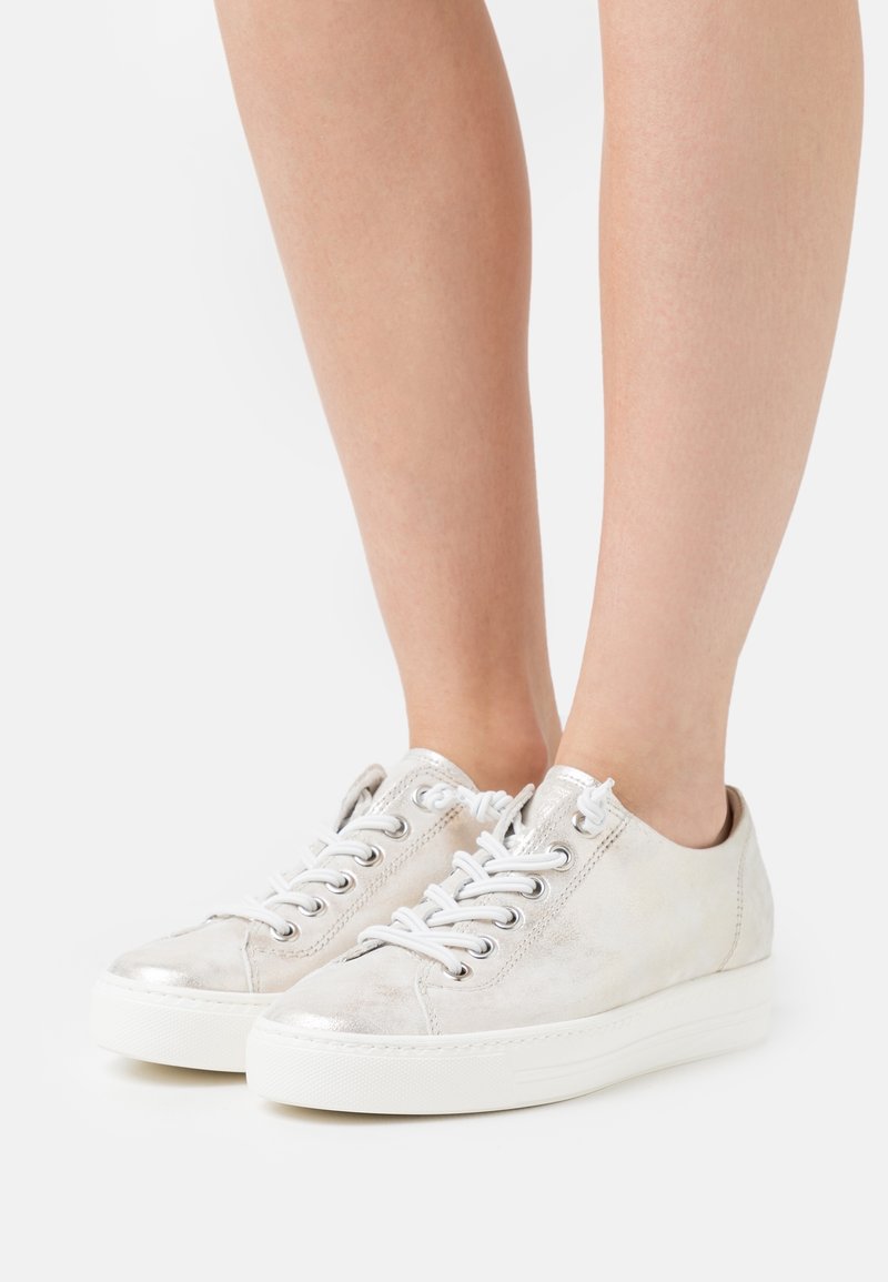Jambes portant des baskets basses argentées métalliques à lacets avec des semelles épaisses en caoutchouc blanc, présentées sur fond blanc.