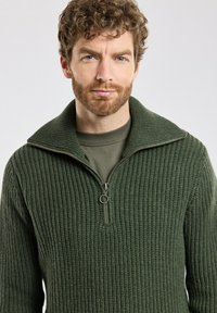 Pull à zip vert foncé avec une texture côtelée et un col montant, superposé sur une chemise verte assortie. Détails de couture subtils.