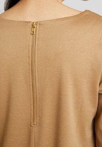 Garbato di colore beige con scollatura rotonda e una zip dorata visibile sul retro, caratterizzato da una texture di tessuto liscia.