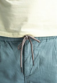 Lichtgele katoenen t-shirt gecombineerd met tealkleurige shorts met trekkoord. De shorts hebben een geknoopt grijs trekkoord met plastic uiteinden.