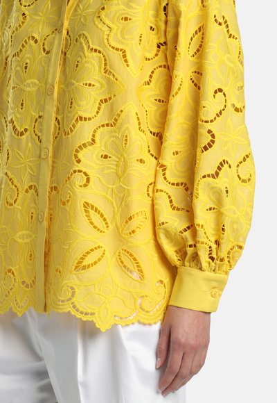 Dzeltena mežģīņu blūze ar ziedu izšuvumu izgriezumiem, pogotu priekšpusi, balonveida piedurknēm un izgrieztu apakšu, valkāta kopā ar baltām biksēm.