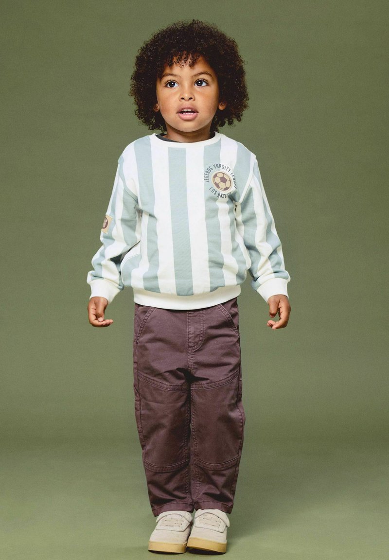 Jeune enfant aux cheveux bouclés portant une chemise rayée à manches longues, un pantalon marron et des chaussures blanches, debout devant un fond vert uni.