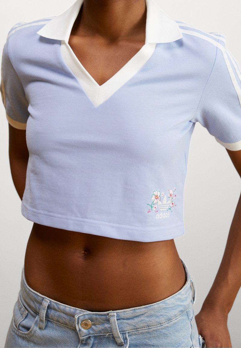 Polo shirt di un blu chiaro tagliato, con colletto bianco, caratterizzato da tre righe bianche sulle maniche e ricami floreali sul lato sinistro.