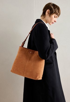 Beige suède totebag met een gladde textuur, voorzien van een bruine leren schouderband en een ronde logo-accent aan de voorkant.