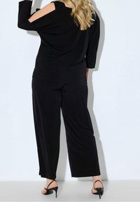 Blonde Frau trägt ein schwarzes Cold-Shoulder-Top und weite Hosen, steht mit dem Rücken zur Kamera, in schwarzen High Heels.