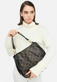 Borsa a tracolla nera e marrone con motivo e accessori dorati, mostrata nelle mani di una persona su uno sfondo chiaro.