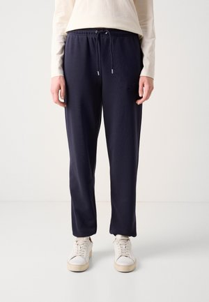 Person wearing navy blue drawstring sweatpants, white long-sleeve top, and white sneakers against a plain light background.