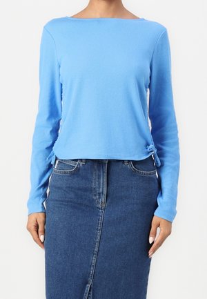 Kobieta ubrana w jasnoniebieską prążkowaną bluzkę z długim rękawem i troczkami po bokach oraz wysoką spódnicę z niebieskiego denimu z przednim rozcięciem.