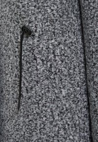 Grijze fleece stof met een gestructureerd, pluizig oppervlak. Voorzien van een trekkoord met een zwarte stopper voor aanpassing.
