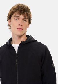 Schwarze Zip-Hoodie mit gerippter Textur, einer Kapuze und dezenten Akzenten. Verfügt über einen Reißverschluss und ein Logopatch auf dem Ärmel.