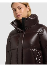 Kvinna i en mörkbrun glänsande vadderad jacka med hög krage, stående mot en enfärgad ljus bakgrund.