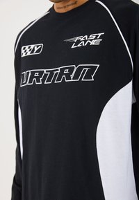 Long-sleeve black shirt with white accents; features bold white text, graphic prints, and a sporty design. Soft fabric with smooth texture.