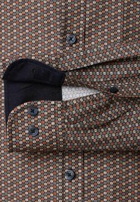 Geknoopte blouse met een veelkleurig geometrisch patroon in bruin, oranje en crème. Heeft een donkere kraag en contrasterende manchetten, met zichtbare stofstructuur.