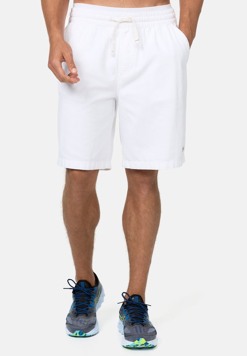 Pantalones cortos de algodón blancos con una cinturilla elástica y cordón, que cuentan con bolsillos laterales y un dobladillo enrollado. Combinados con zapatillas deportivas en gris y azul.