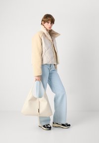 Jachetă pufoasă de crem și bej cu guler din blană de miel, blugi blue deschis și o geantă mare de crem. Tenisi negri și crem completează look-ul.