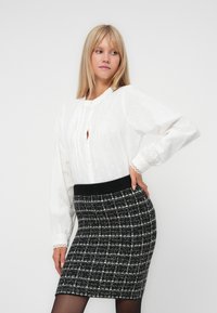 Blondýnka v bílé blůze s dlouhým rukávem a černo-bílé kostkované sukni pózuje s rukou na boku na jednobarevném pozadí.