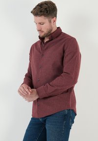 Camisa de botones color marrón con una textura en la tela, de mangas largas y color de cuello estándar. Presenta botones oscuros y un bajo curvado. Llevada con vaqueros azules.